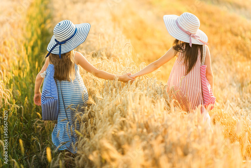 Two girls walk on a rye field in linen dresses and hats. Fashion, style, summer children's clothing. Looking from the back. photo