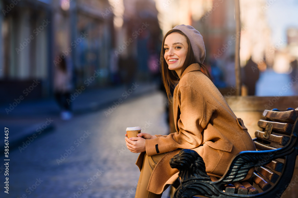 Young smiling woman girl drinking morning coffee resting on bench. Old town street background. Warm up with coffee in cold weather. Young smiling woman girl drinking morning coffee resting on bench. Old town street background. Warm up with coffee in cold weather.