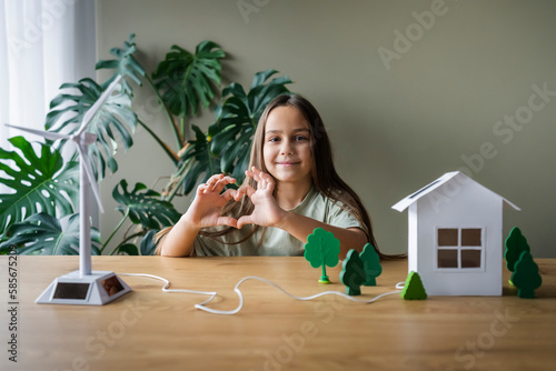 Girl making heart gesture sitting at table with with wind turbine model