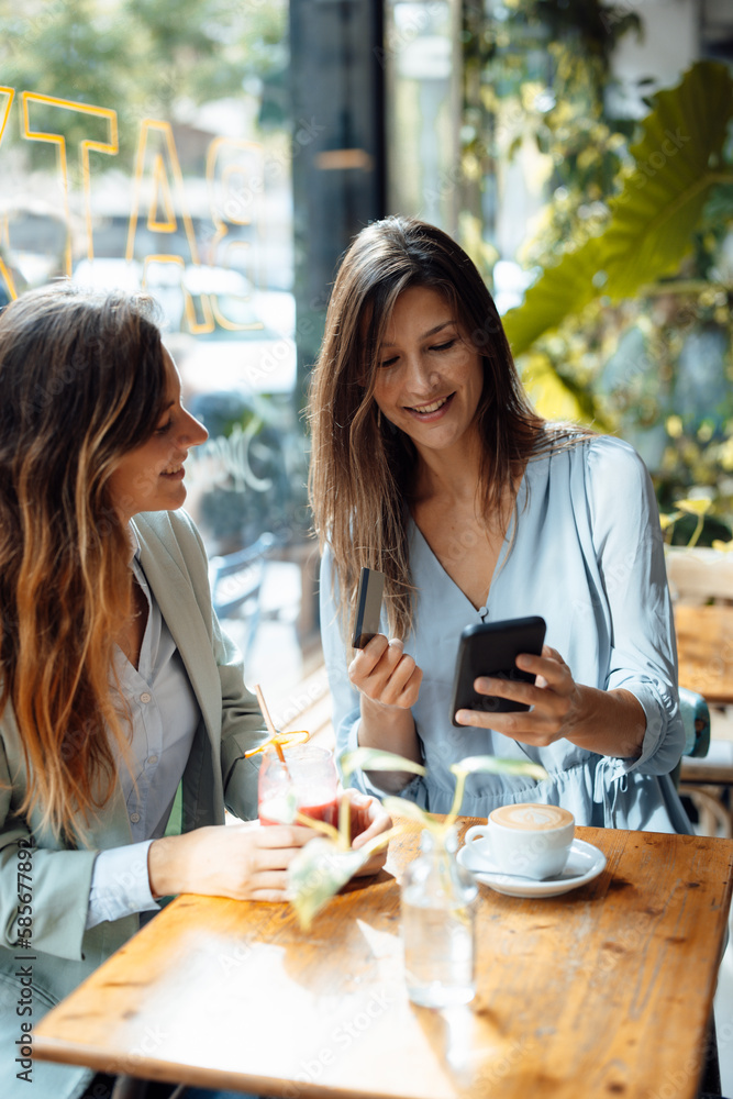 Woman with friend holding credit card using smart phone in cafe Woman with friend holding credit card using smart phone in cafe