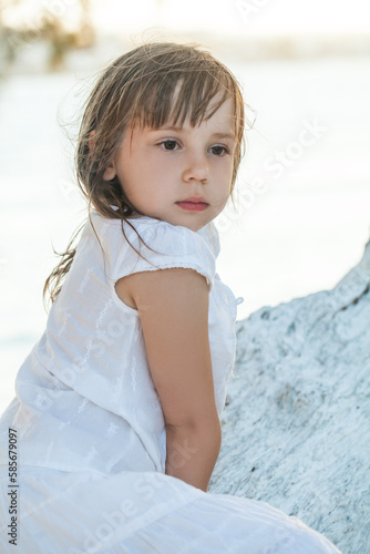 Portrait of child girl in white clothes sitting near the tree on background of the blue sea