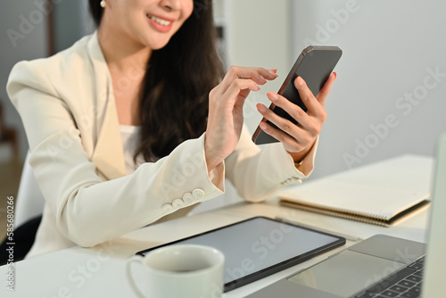 Smiling young female worker sitting at workplace chatting with friends, reading text message on mobile phone
