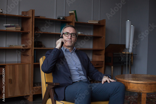 Portrait of elderly handsome successful businessman in suit and glasses talking on his mobile phone on grey office wall background with bookshelf