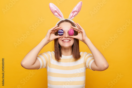 Portrait of cheerful funny woman wearing bunny ears covering eyes with easter eggs, having fun, smiling to camera, isolated over yellow background, hiding face. © sementsova321