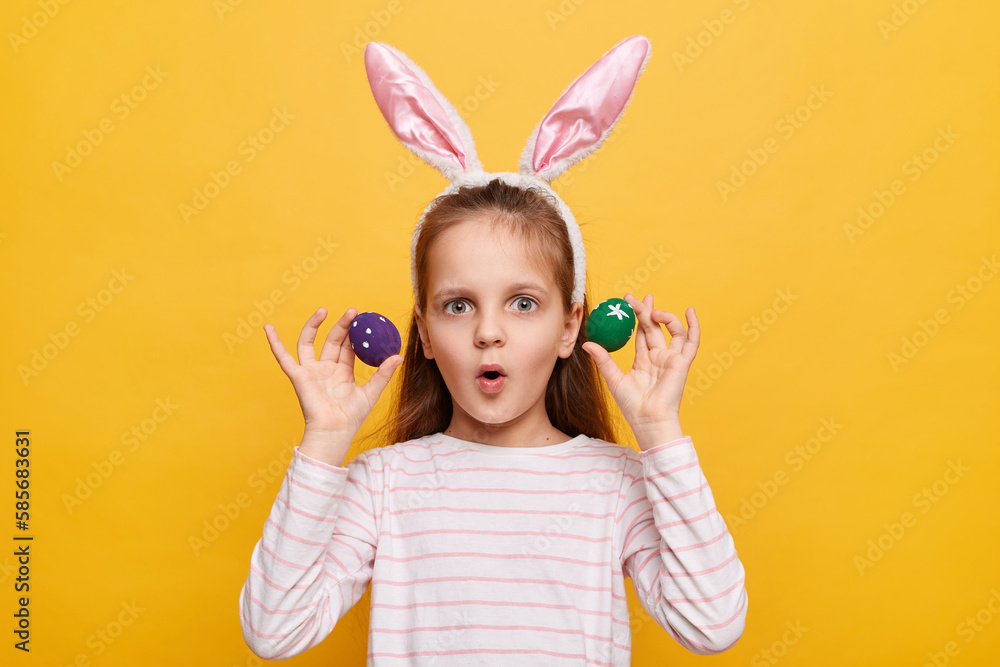 Indoor shot of shocked surprised girl with rabbit ears on her head with eggs isolated on yellow background, looking at camera with big eyes, sees something astonishing. Indoor shot of shocked surprised girl with rabbit ears on her head with eggs isolated on yellow background, looking at camera with big eyes, sees something astonishing.