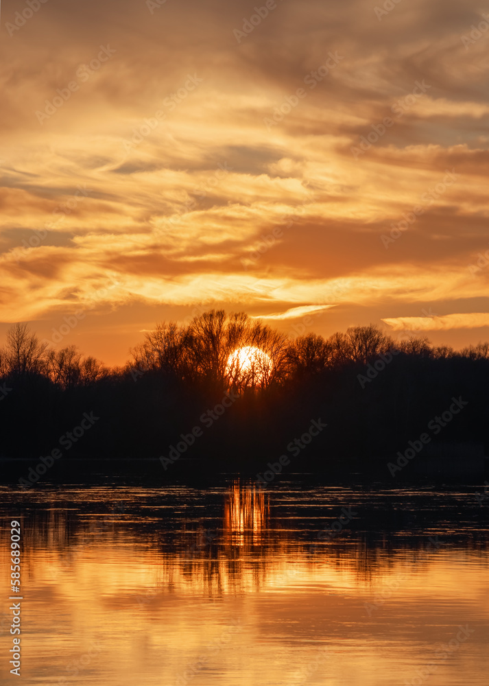 Scenic orange sunset with clouds over forest and river. The Sun hides behind the trees Scenic orange sunset with clouds over forest and river. The Sun hides behind the trees