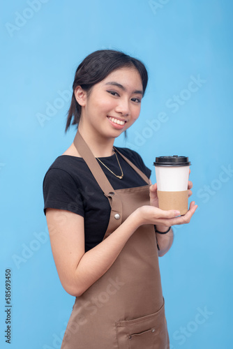 A young and lovely asian female barista presenting a cup of coffee. Wearing a brown apron and black shirt and against a light blue background.