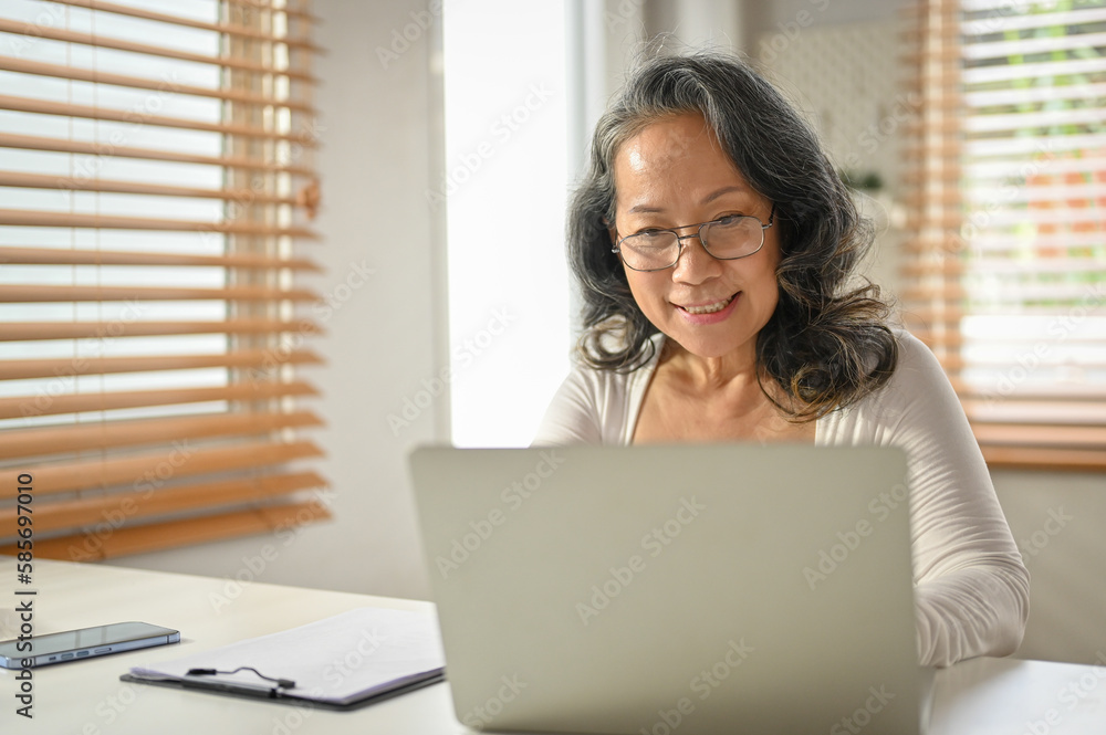 Smiling 60-year-old Asian businesswoman working on her business tasks on a laptop Smiling 60-year-old Asian businesswoman working on her business tasks on a laptop