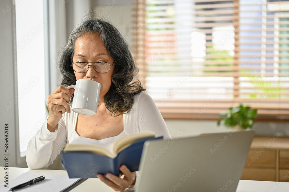 Relaxed and calm 60s Asian-aged woman sipping her morning coffee and reading a book Relaxed and calm 60s Asian-aged woman sipping her morning coffee and reading a book