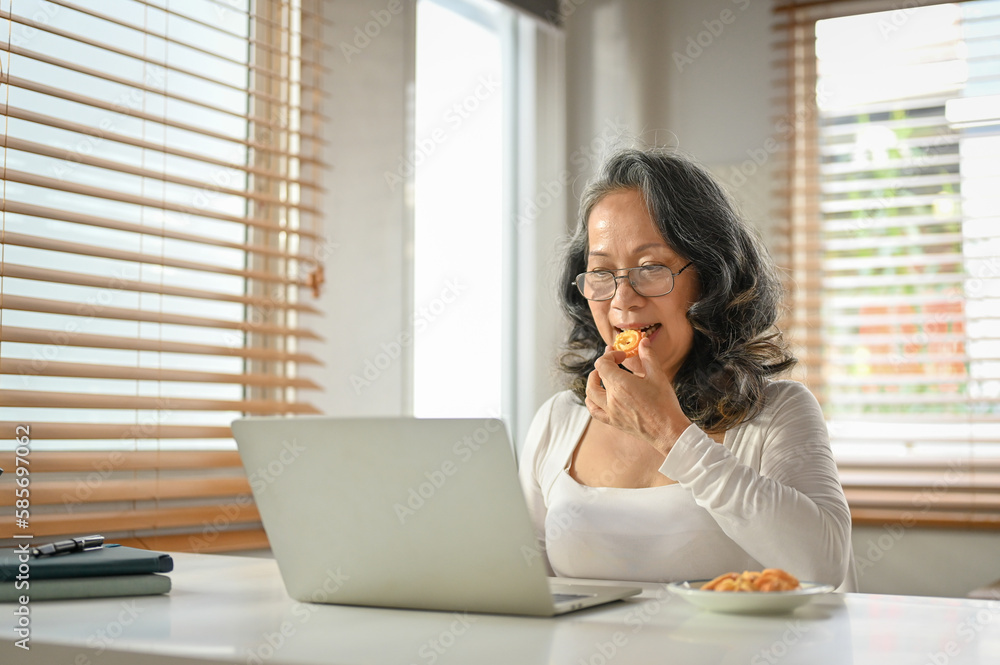 Chilling 60s Asian-aged woman eating cookies while using her laptop at her home workspace. Chilling 60s Asian-aged woman eating cookies while using her laptop at her home workspace.
