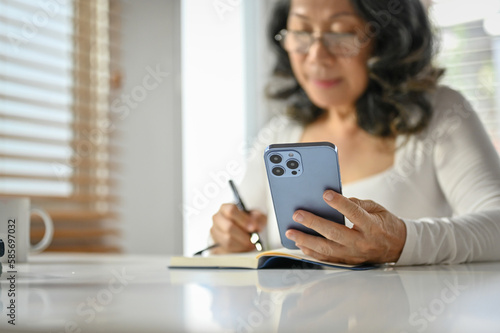 Selective focus, An Asian-aged woman using her phone and making lists on her notepad