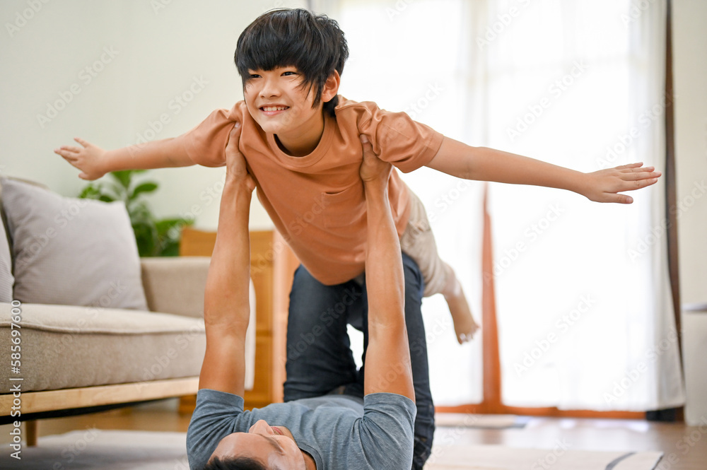 Fototapeta premium Loving Asian dad lying on the living room floor playing airplane with little son