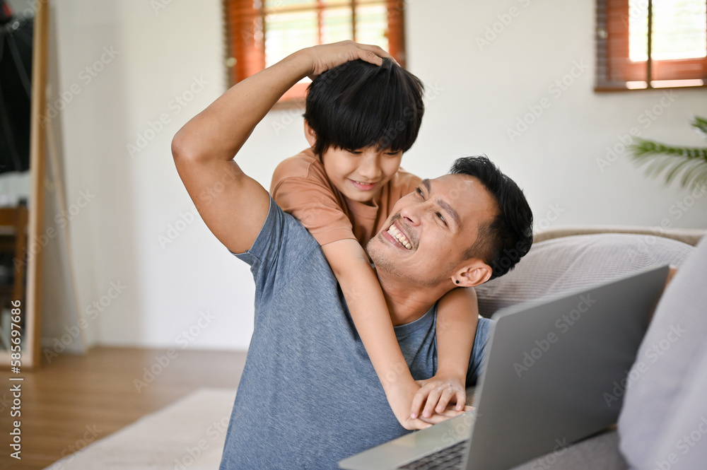 Cute little Asian boy hugging his father from behind while he works in the living room. 