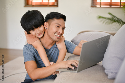 Cute little Asian boy hugging his father from behind while he works on his laptop