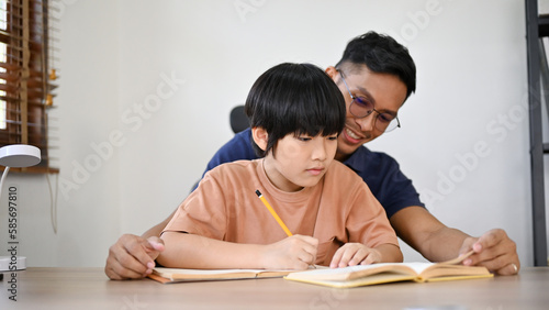 Cute Asian little boy focusing on his homework while his dad is teaching and helping.