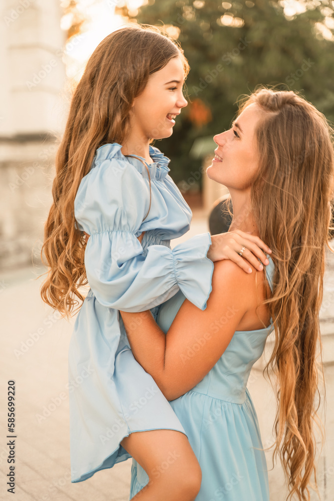 Portrait of a mother and daughter in blue dresses with flowing long hair against the backdrop of a sunset and a white building. They look at each other. Family stories on the weekend Portrait of a mother and daughter in blue dresses with flowing long hair against the backdrop of a sunset and a white building. They look at each other. Family stories on the weekend