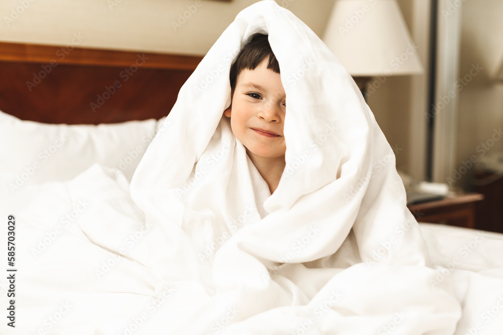 Little cute boy wrapped himself in a white blanket while sitting on the bed and smiling. Happy kid look surprised, his hands on chin, amazement toddler boy. Good morning. Little cute boy wrapped himself in a white blanket while sitting on the bed and smiling. Happy kid look surprised, his hands on chin, amazement toddler boy. Good morning.