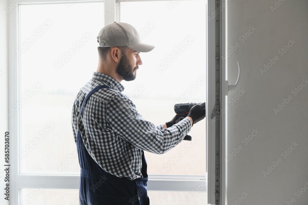 Workman in overalls installing or adjusting plastic windows in the living room at home Workman in overalls installing or adjusting plastic windows in the living room at home