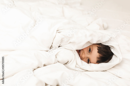 Little cute boy wrapped himself in a white blanket while lying on the bed and smiling. Happy kid look happy, amazement toddler boy. Good morning.