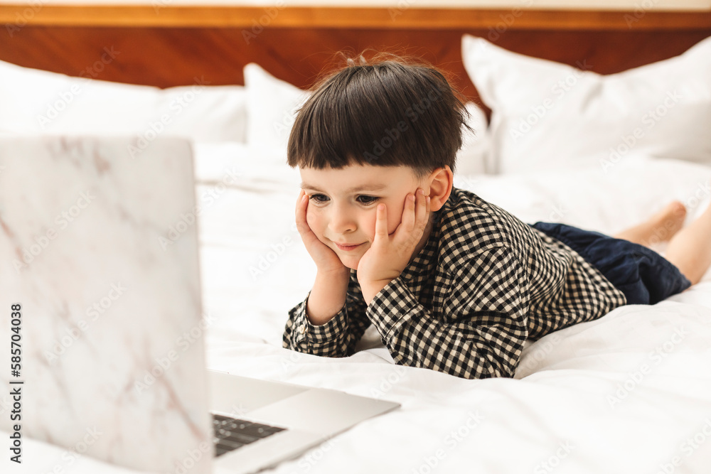 Smart looking preschool boy lying on the bed using notebook computer in his room hold his head and looking at the screen. Online learning, Home preschool, New normal, Home Based Learning Smart looking preschool boy lying on the bed using notebook computer in his room hold his head and looking at the screen. Online learning, Home preschool, New normal, Home Based Learning