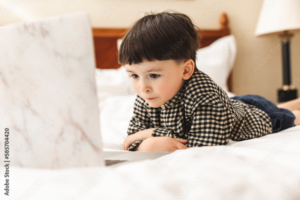 Smart looking preschool boy lying on the bed using notebook computer in his room looking at the screen. Online learning, Home preschool, New normal, Home Based Learning Smart looking preschool boy lying on the bed using notebook computer in his room looking at the screen. Online learning, Home preschool, New normal, Home Based Learning