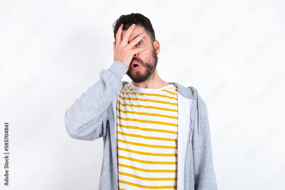Young caucasian mán wearing trendy clothes over white background peeking in shock covering face and eyes with hand, looking through fingers with embarrassed expression. Young caucasian mán wearing trendy clothes over white background peeking in shock covering face and eyes with hand, looking through fingers with embarrassed expression.