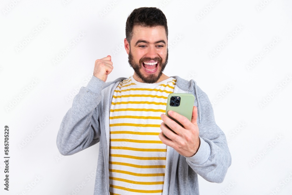 Young caucasian mán wearing trendy clothes over white background holding in hands cell and rising his fist up being excited after reading good news. Young caucasian mán wearing trendy clothes over white background holding in hands cell and rising his fist up being excited after reading good news.
