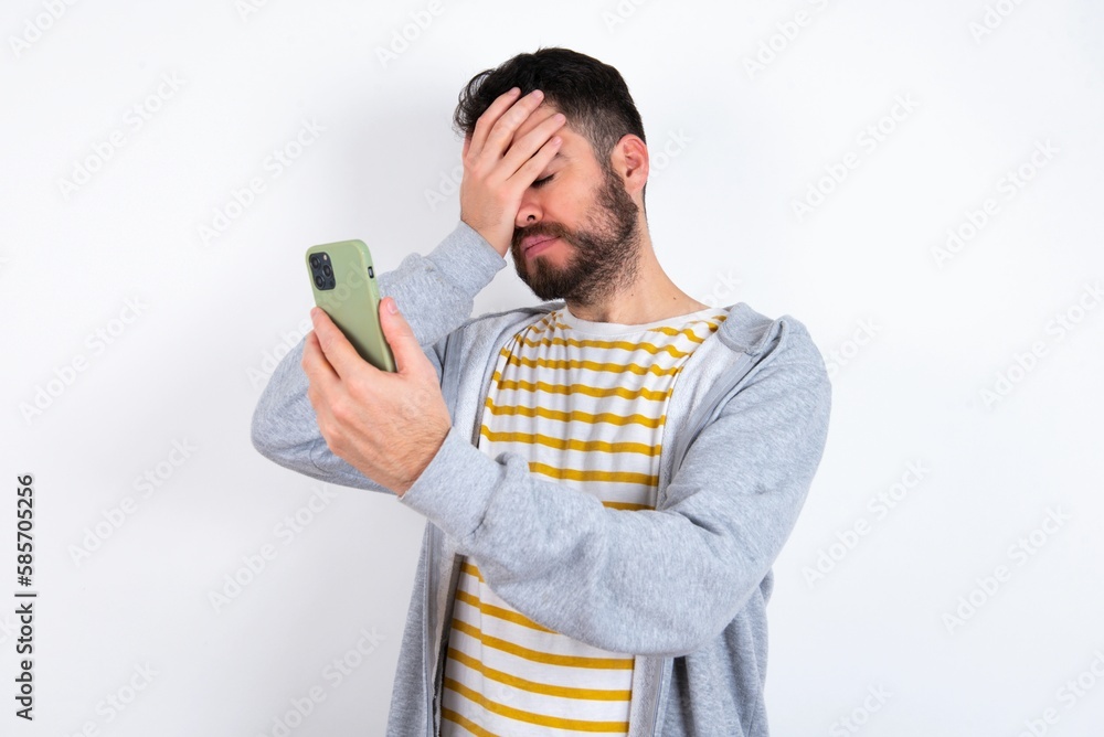 Young caucasian mán wearing trendy clothes over white background looking at smart phone feeling sad holding hand on face. Young caucasian mán wearing trendy clothes over white background looking at smart phone feeling sad holding hand on face.