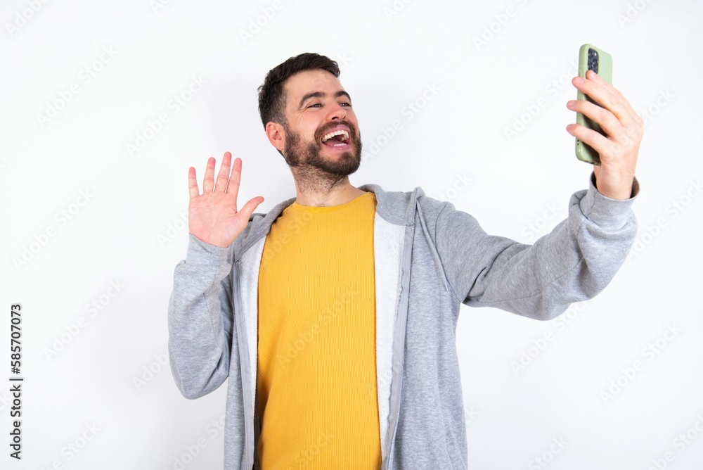 Φωτογραφία Young caucasian mán wearing trendy clothes over white background holds modern mobile phone and makes video call waves palm in hello gesture