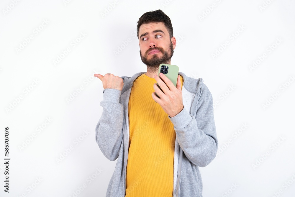 Young caucasian mán wearing trendy clothes over white background points thumb away and shows blank space aside, holds mobile phone for sending text messages. Young caucasian mán wearing trendy clothes over white background points thumb away and shows blank space aside, holds mobile phone for sending text messages.