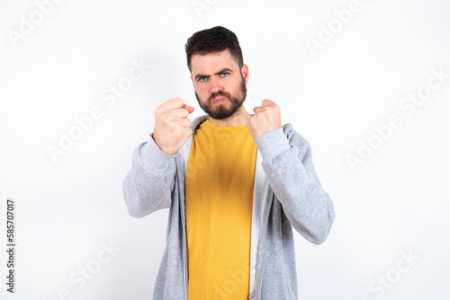 Displeased annoyed Young caucasian mán wearing trendy clothes over white wall clenches fists, gestures pissed, ready to revenge, looks with aggression at camera stands full of hate, being pressured