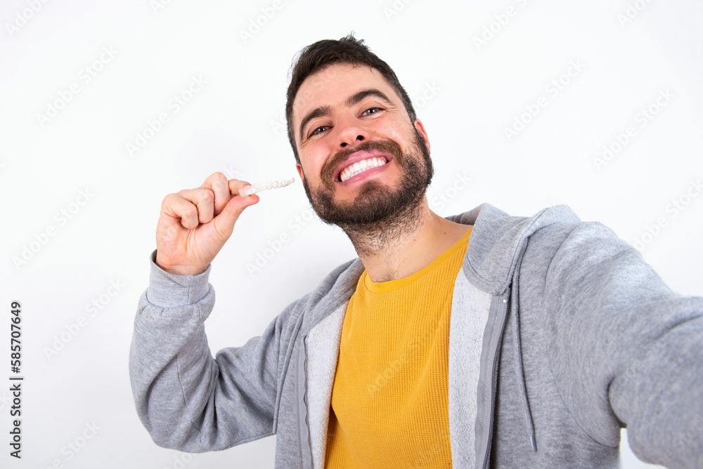 Young caucasian mán wearing trendy clothes over white background make selfie holding an invisible braces aligner, recommending this new treatment. Dental healthcare concept. Young caucasian mán wearing trendy clothes over white background make selfie holding an invisible braces aligner, recommending this new treatment. Dental healthcare concept.
