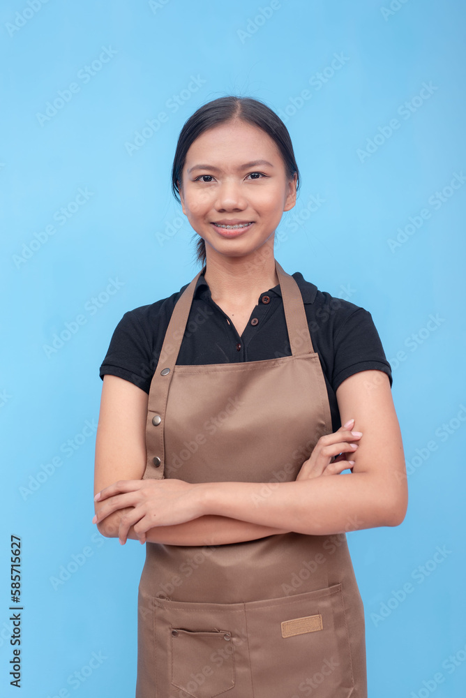 A typical female asian barista or waitress with arms crossed and wearing a brown apron and black shirt and hair tied up. Against a light blue background. A typical female asian barista or waitress with arms crossed and wearing a brown apron and black shirt and hair tied up. Against a light blue background.