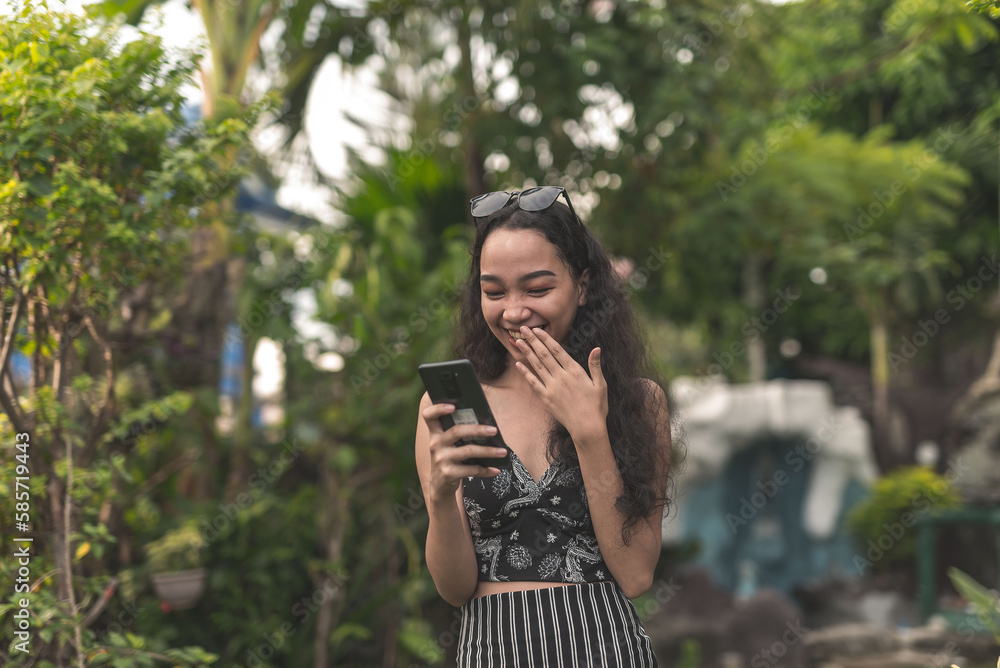 A pretty young Filipina woman with curly hair wearing a nice black top and long skirt blushes in surprise while chatting on her phone at the garden. A pretty young Filipina woman with curly hair wearing a nice black top and long skirt blushes in surprise while chatting on her phone at the garden.