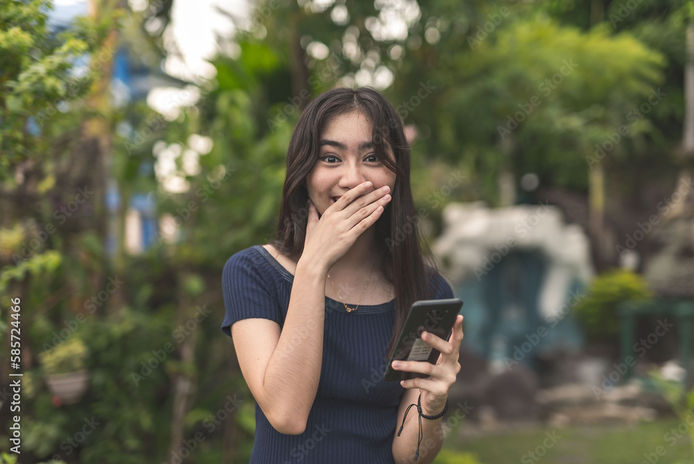 A pretty young woman reacts with excitement after checking the latest rumors on social media on her phone at the garden. A pretty young woman reacts with excitement after checking the latest rumors on social media on her phone at the garden.