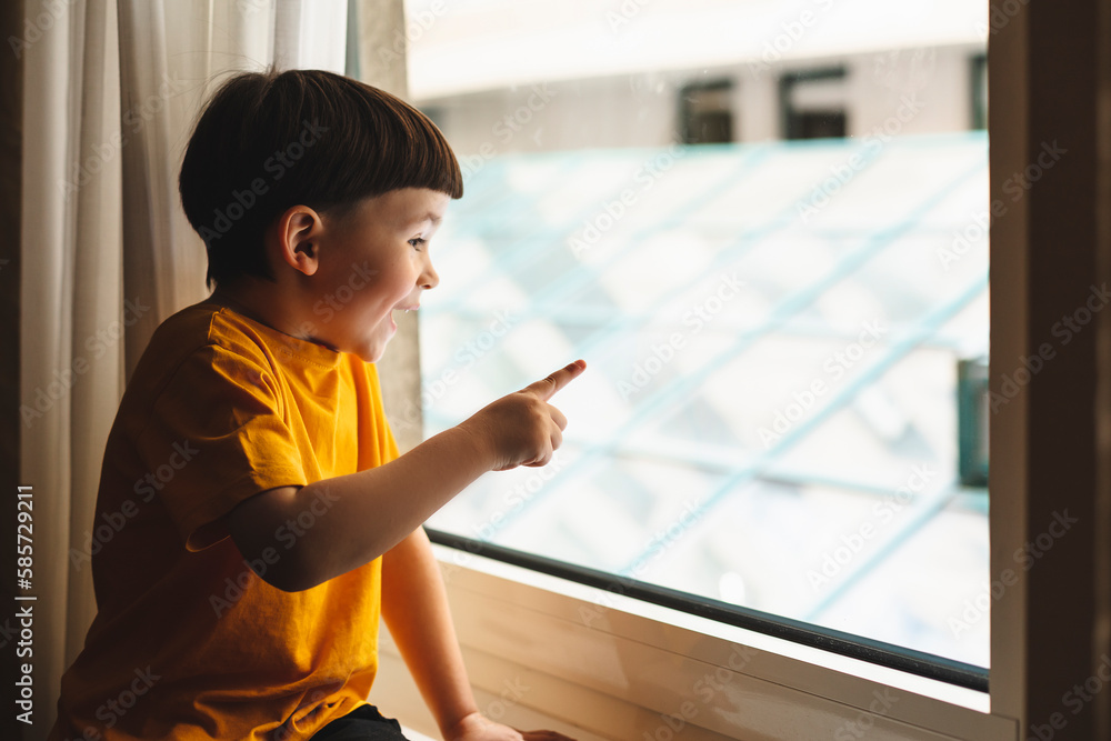 Little cute attractive boy sitting on windowsill and look at window, boy point his finger on something, what he saw on window. Kid look shocked amazement and surprised, he wear yellow t-shirt at home. Little cute attractive boy sitting on windowsill and look at window, boy point his finger on something, what he saw on window. Kid look shocked amazement and surprised, he wear yellow t-shirt at home.