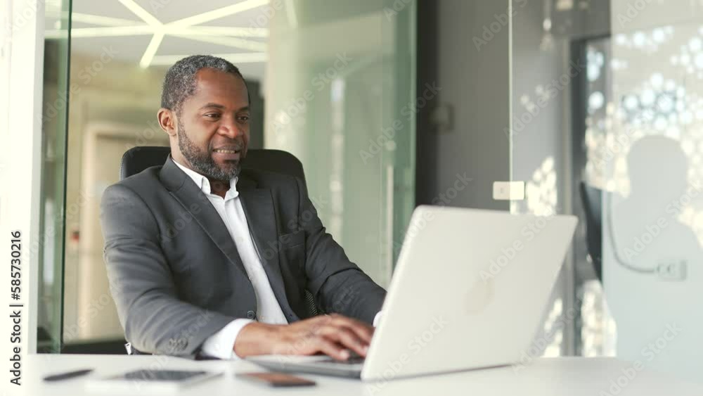 Excited mature african american man in formal suit watching sports match while sitting at workplace using laptop in office Happy excited businessman cheering for favorite team with emotional gesturing Excited mature african american man in formal suit watching sports match while sitting at workplace using laptop in office Happy excited businessman cheering for favorite team with emotional gesturing