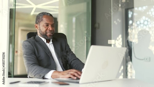 Excited mature african american man in formal suit watching sports match while sitting at workplace using laptop in office Happy excited businessman cheering for favorite team with emotional gesturing photo