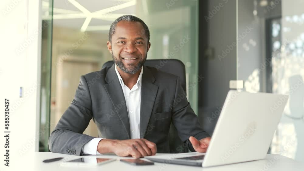 Portrait of a confident mature african american man in a formal suit sitting at a workplace using a laptop in office. Happy smiling middle aged businessman looking at the camera with a friendly look Portrait of a confident mature african american man in a formal suit sitting at a workplace using a laptop in office. Happy smiling middle aged businessman looking at the camera with a friendly look