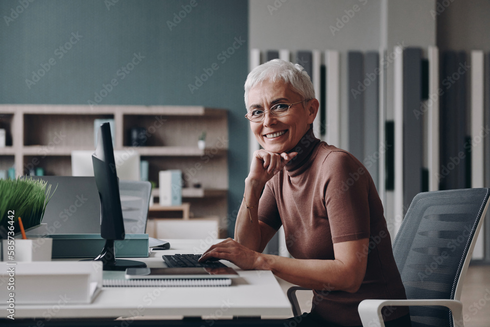 Senior businesswoman looking at camera and smiling while sitting at her working place in office Senior businesswoman looking at camera and smiling while sitting at her working place in office