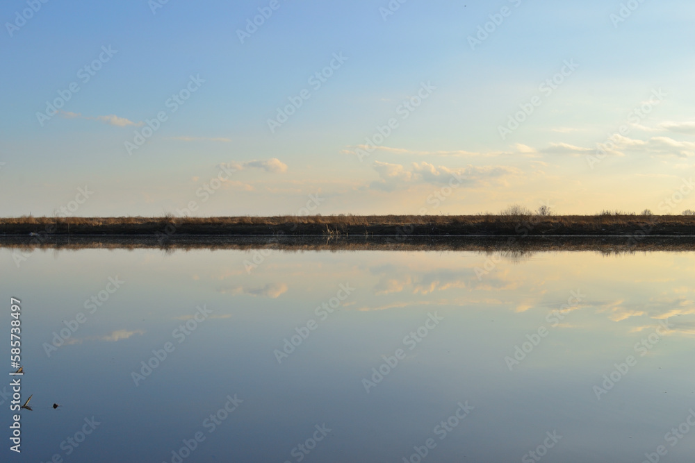 Meadow bank of the river, going beyond the horizon, divide the photo in half. Reflection of the sky in the water of the river. Sunset light.