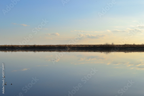 Meadow bank of the river  going beyond the horizon  divide the photo in half. Reflection of the sky in the water of the river. Sunset light.