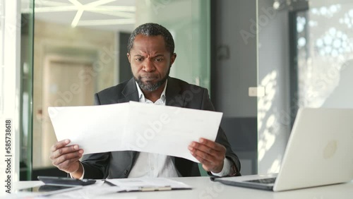 Confused mature businessman having difficulty with paper work sitting at workplace in modern office. The accountant looks at the documents, spreads his hands and cannot understand the problem
