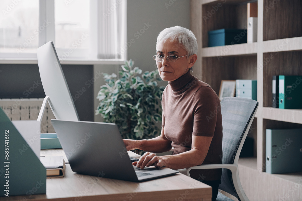 Confident senior businesswoman working at two computers while sitting in the office Confident senior businesswoman working at two computers while sitting in the office