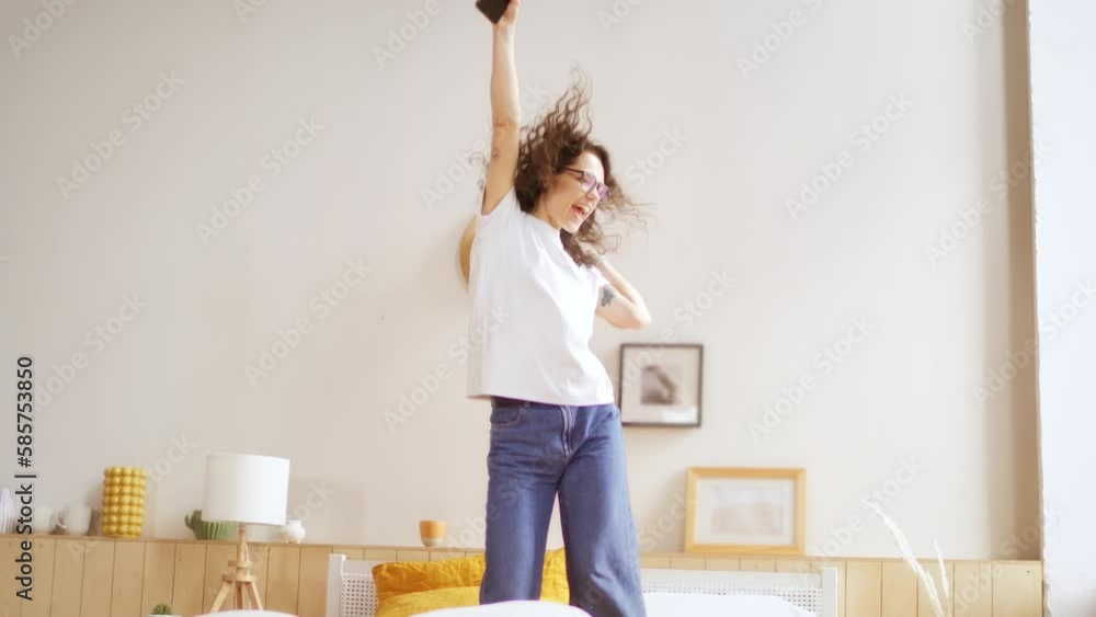 Excited young woman winner looks at mobile phone celebrates online success at home. Girl enjoys good news and jumps on the bed with happiness. lady gets new distance job opportunity. Excited young woman winner looks at mobile phone celebrates online success at home. Girl enjoys good news and jumps on the bed with happiness. lady gets new distance job opportunity.