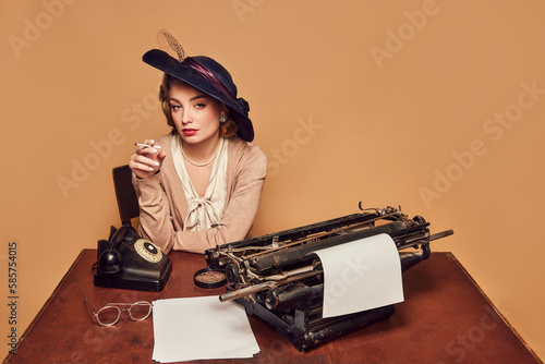 Attractive woman writer wearing old-fashioned clothes sitting at table and smoking over beige background