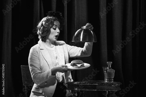 Monochrome portrait of adorable woman wearing vintage costume holding burger with surprised face and open mouth photo