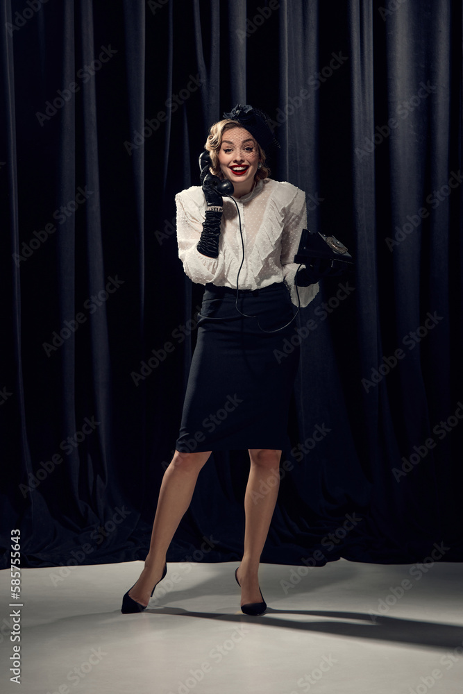 Pretty woman wearing old-fashioned clothes holding phone and talking with pleasure over dark background