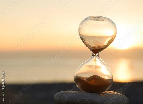 Hourglass on the beach, on the Black sea coast. Hourglass close up in a warm golden morning sunlight on a pebble beach starting time for a new day or running of time with side copy space.