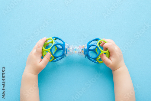 Baby boy hands playing with colorful rattle dumbbell on light blue table background. Pastel color. Closeup. Point of view shot. Toy of development for infant. Top down view. photo